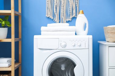 İçerideki mavi duvarın yanındaki çamaşır makinesinin üzerinde deterjan olan temiz havlu ve şişe yığını.