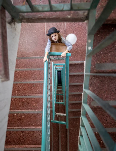 一个戴着帽子 头戴白色气球的漂亮姑娘站在一座大楼的楼梯上抬头看 — 图库照片