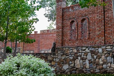 Tamamlanmamış kırmızı tuğla, eski ve desenli taş. Pencereler için geniş oval açıklıklar, açık gökyüzünün altında ağaçlar ve yeşillikler arasında