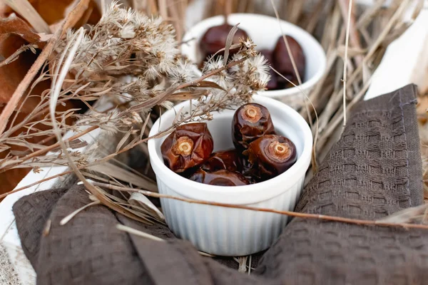 Dried dates, sweet fruits of the date palm in small ceramic forms in a wooden white box, decorated with dry leaves and yellow grass, various autumn twigs. Composition on a stool, late autumn