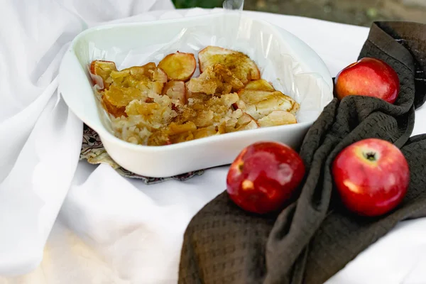 Collapsed soft baked apples in a white ceramic square shape on a special package. A white tablecloth is laid carelessly on the table, next to a dark napkin and ripe juicy red apples