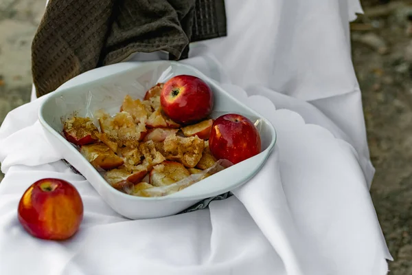 Collapsed soft baked apples in a white ceramic square shape on a special package. A white tablecloth is laid carelessly on the table, next to a dark napkin and ripe juicy red apples