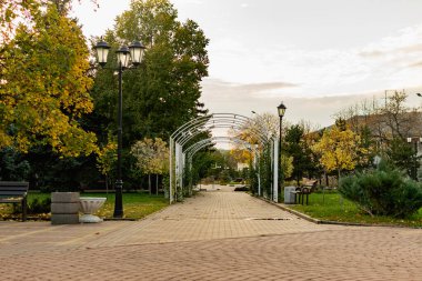 Şehrin halka açık parkında yürüyüş. Güzel beyaz metal kemer, bir sürü yeşillik, ağaçlar ve kozalaklı ağaçlar, kenar boyunca çimenler, banklar ve fenerler. Yol fayanslıdır.