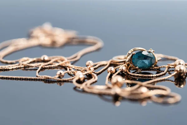A gold chain with a pendant  and a Topaz ring lie on a dark reflective surface