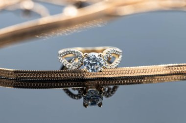 Yılan derisi örgülü altın bir zincir ve büyük bir elmas yüzük beyaz yüzeye yansıyor.