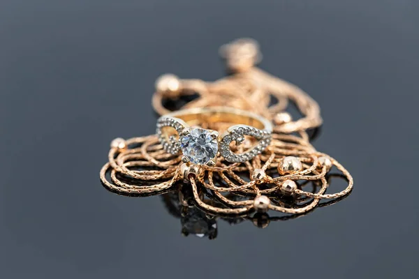 A gold chain with gold beads weave and a large diamond ring are reflected on the white surface