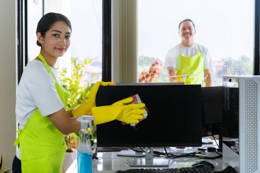 Çevre dostu ofis temizliği hizmeti sunan önlüklü profesyonel temizlikçi çift, sağlıklı bir iş yeri için pencere, bilgisayar ve toplantı odalarını sterilize ederken yeşil ürünler kullanıyor.