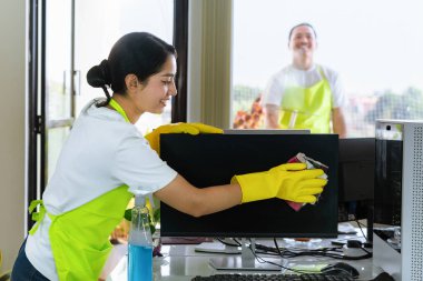 Çevre dostu ofis temizliği hizmeti sunan önlüklü profesyonel temizlikçi çift, sağlıklı bir iş yeri için pencere, bilgisayar ve toplantı odalarını sterilize ederken yeşil ürünler kullanıyor.