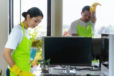 Çevre dostu ofis temizliği hizmeti sunan önlüklü profesyonel temizlikçi çift, sağlıklı bir iş yeri için pencere, bilgisayar ve toplantı odalarını sterilize ederken yeşil ürünler kullanıyor.