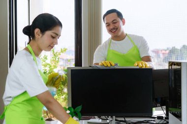 Çevre dostu ofis temizliği hizmeti sunan önlüklü profesyonel temizlikçi çift, sağlıklı bir iş yeri için pencere, bilgisayar ve toplantı odalarını sterilize ederken yeşil ürünler kullanıyor.