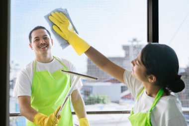 Çevre dostu ofis temizliği hizmeti sunan önlüklü profesyonel temizlikçi çift, sağlıklı bir iş yeri için pencere, bilgisayar ve toplantı odalarını sterilize ederken yeşil ürünler kullanıyor.