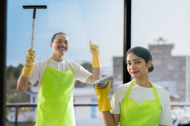 Çevre dostu ofis temizliği hizmeti sunan önlüklü profesyonel temizlikçi çift, sağlıklı bir iş yeri için pencere, bilgisayar ve toplantı odalarını sterilize ederken yeşil ürünler kullanıyor.