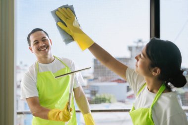 Çevre dostu ofis temizliği hizmeti sunan önlüklü profesyonel temizlikçi çift, sağlıklı bir iş yeri için pencere, bilgisayar ve toplantı odalarını sterilize ederken yeşil ürünler kullanıyor.