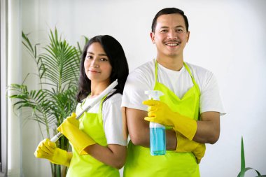 Çevre dostu ofis temizliği hizmeti sunan önlüklü profesyonel temizlikçi çift, sağlıklı bir iş yeri için pencere, bilgisayar ve toplantı odalarını sterilize ederken yeşil ürünler kullanıyor.