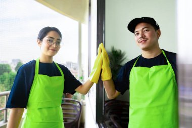 Çevre dostu ofis temizliği hizmeti sunan önlüklü profesyonel temizlikçi çift, sağlıklı bir iş yeri için pencere, bilgisayar ve toplantı odalarını sterilize ederken yeşil ürünler kullanıyor.