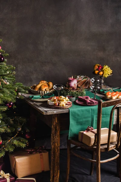 Stílusos asztal beállítása karácsonyi családi vacsora karácsonyfa a háttérben, és ajándékok a széken. — Stock Fotó