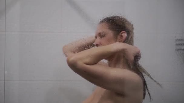Frau wäscht Haare hinter Glas der Duschkabine — Stockvideo