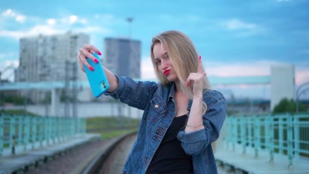 En ung blondine på skinnene tar en selfie på en smarttelefon.. – stockvideo