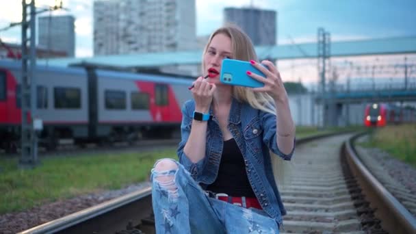 Una giovane donna, bionda, sui binari della ferrovia, si fa un selfie su uno smartphone. — Video Stock