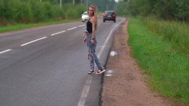 Bir kız otobanda yürüyor ve geçen bir arabayı durdurmaya çalışıyor.