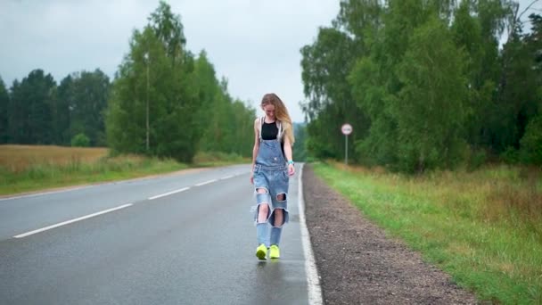 Seorang gadis muda berambut pirang berjalan di sepanjang jalan raya dalam cuaca mendung — Stok Video