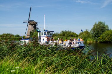Kinderdijk, Hollanda, Ağustos 2019. Hollanda kırsalında, uzun yeşil otlarla kaplı kanallarla çevrili, mükemmel durumdaki tarihi bir yel değirmeni. Bir turist teknesi geçiyor..