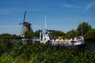 Kinderdijk, Hollanda, Ağustos 2019. Hollanda kırsalında, uzun yeşil otlarla kaplı kanallarla çevrili, mükemmel durumdaki tarihi bir yel değirmeni. Bir turist teknesi geçiyor..