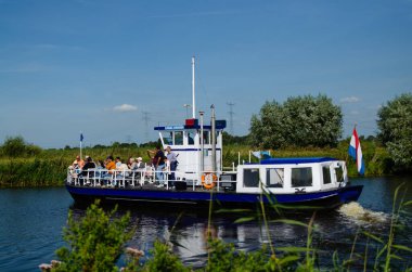 Kinderdijk, Hollanda, Ağustos 2019. Bir gezi teknesi, bölgenin tarihi yel değirmenlerini ziyaret etmek için Hollanda kırsal kesimindeki kanalları geziyor. Güneşli güzel bir gün, tekne kalabalık..