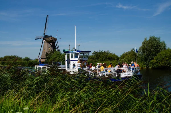 Kinderdijk, Hollanda, Ağustos 2019. Hollanda kırsalında, uzun yeşil otlarla kaplı kanallarla çevrili, mükemmel durumdaki tarihi bir yel değirmeni. Bir turist teknesi geçiyor..