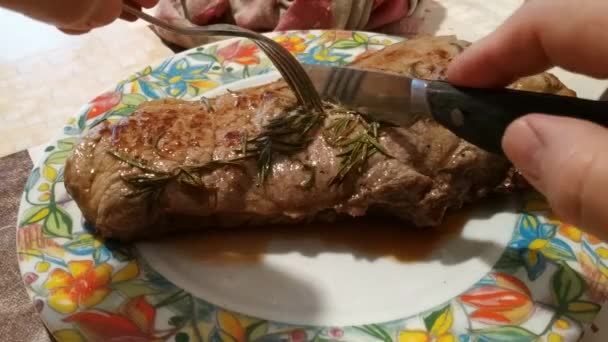 A large beef steak was cooked and seasoned with rosemary. Still smoking in the plate, knife and fork cut it in half. In the background the table is set. 25fps — Stock Video