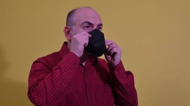 Orta yaşlı beyaz bir adam kendini koronavirüsten korumak için siyah bir maske takıyor. Kişisel bakım konsepti. Stüdyo arkaplanı sarı renkte.