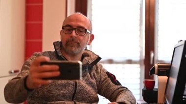 Orta yaşlı, beyaz bir adam çalışma masasında oturuyor. Ev mutfağında. Selfie çekiyor. Evden ve teknoloji konseptinden çalışma.