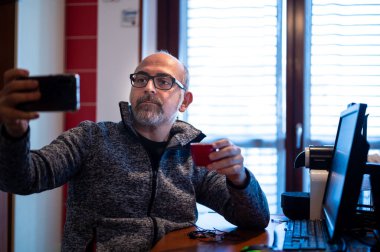 Orta yaşlı, beyaz bir adam çalışma masasında oturuyor. Ev mutfağında. Selfie çekiyor. Evden ve teknoloji konseptinden çalışma.