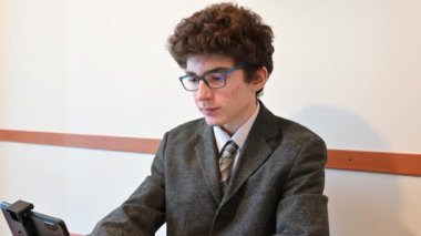 Genç beyaz bir çocuk muhabir gibi giyinmiş ve uzaktan röportaj yapmaya hazırlanıyor. Küçük bir tripodun desteklediği akıllı telefonuyla test ediyor. Heyecanlı..