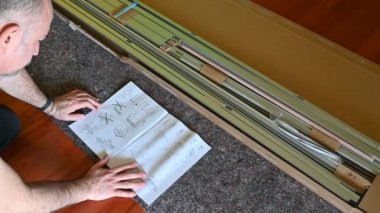 Ev işi: bir Ikea mobilyası monte etmek. Beyaz adamın eli, el kitabının sayfalarını dikkatle karıştırıyor. El kitabının altında yerleşmiş mobilyaların parçaları var..