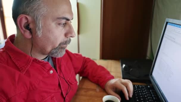 自宅で働く中年の白人男性の肖像 彼は自宅の部屋で作業ステーションを作成しています パソコンを操作しながら白いカップからコーヒーを飲む 黒いイヤホンを持ってる — ストック動画