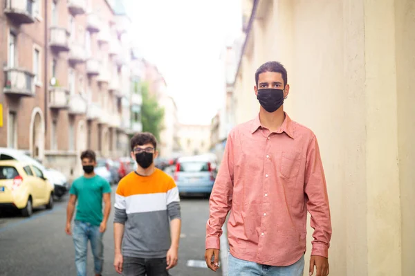 Group of friends walking on the city street with black protective mask. Pandemic time. New concept of normal lifestyle with young people covered with a protective mask. Focus on the guy on the right.