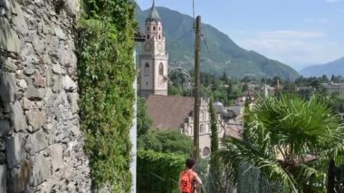 Merano, İtalya, Haziran 2021. Katedral 'in çan kulesinin eğik görüntüleri. Manzaralı yolda bir çocuk şehrin büyüleyici manzarasını izliyor..