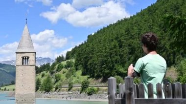 Curon, İtalya, Haziran 2021. Curon 'un (Graun) eski çan kulesinin güzel yaz günündeki görüntüleri Resia Gölü' nün sularından yükseliyor. Yeşil tişörtlü bir adam akıllı telefonuyla fotoğraf çekiyor..