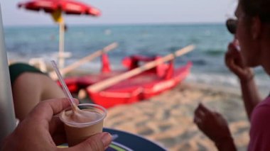 Ön planda bir bardak kremalı kavramsal görüntü, onu yiyen orta yaşlı bir kadını bulanıklaştırdı. Şemsiyeli ve cankurtaran botlu sahilin daha uzağında. Rahatlayın ve tatile çıkın.