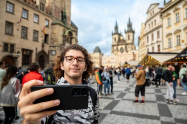 Prag, Çek Cumhuriyeti, 7 Ağustos 2023. Kafkasyalı genç bir adam cep telefonuyla eski kasaba meydanında selfie çekiyor. Teknolojiyi kullanarak. Bulanık arkaplan, seyahat hedefleri.
