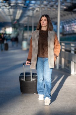 Genç bir kadın, parlak, modern bir havaalanı ya da tren istasyonu terminalinde, siyah bir bavul taşıyarak, seyahat, ayrılış ve bağımsızlığı somutlaştırarak güvenle yürüyor.