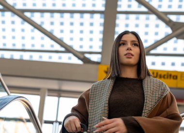 Havalı giyinmiş genç bir kadın, modern bir havaalanı terminalindeki yürüyen merdivende hazır ve kalkış ve seyahat macerasına hazır bir şekilde bavulunu tutarken yukarı bakar.