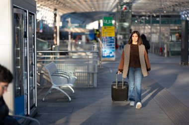 Kadın gezgin, parlak, modern bir tren istasyonu terminalinden tekerlekli bavulu çekiyor, bulanık banliyö sakinleri, şehir içi seyahat yaşam tarzı sahnesinin ortasında kalkışa veya varışa doğru yürüyor