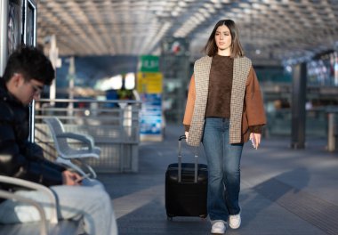 Yuvarlanan bir bavul çeken genç bir kadın, seyahat ederken modern bir havaalanı ya da tren istasyonu terminalinde yürüyor, bulanık arka plan figürleri ve sıcak güneş ışığı sahneyi aydınlatıyor