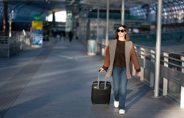 Güneş gözlüklü ve günlük kıyafetli bir kadın, tekerlekli bavulunu parlak bir havaalanı ya da tren istasyonundan geçirir. Gidecekleri yerler arasında güvenli bir şehir seyahatinde tek başına yürür.