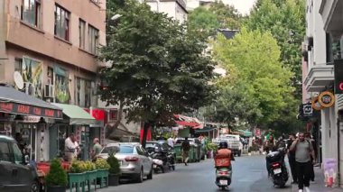 İstanbul, hindi, 5 Ağustos 2025. Yürüyen insanlarla dolu bir şehir caddesi ve scooterlı bir yemek kuryesi. Ticari binalar ve park halindeki araçlar şehir yolunda.