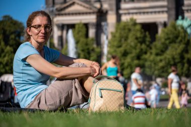 Berlin, Almanya, 11 Ağustos 2023. Yeşil çimlerin üzerinde oturan, dinlenen ve kameraya bakan, arka planda Berlin katedrali olan bir kadın.