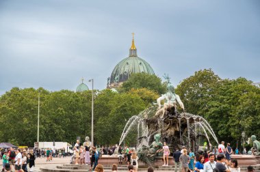 Berlin, Almanya, 11 Ağustos 2023. Neptün çeşmesinin tadını çıkaran insanlar Almanya 'nın başkentinde ağaçların tepesinde yükselen muhteşem berliner dom kubbesiyle