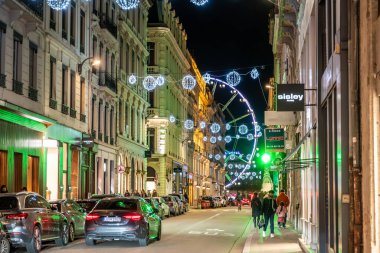 Lyon, Fransa. 27 Aralık 2023. Lyon şehrinin caddesinde şenlikli noel ışıkları, gece dönme dolap, binalar, park edilmiş arabalar ve yayalar sergileniyor.